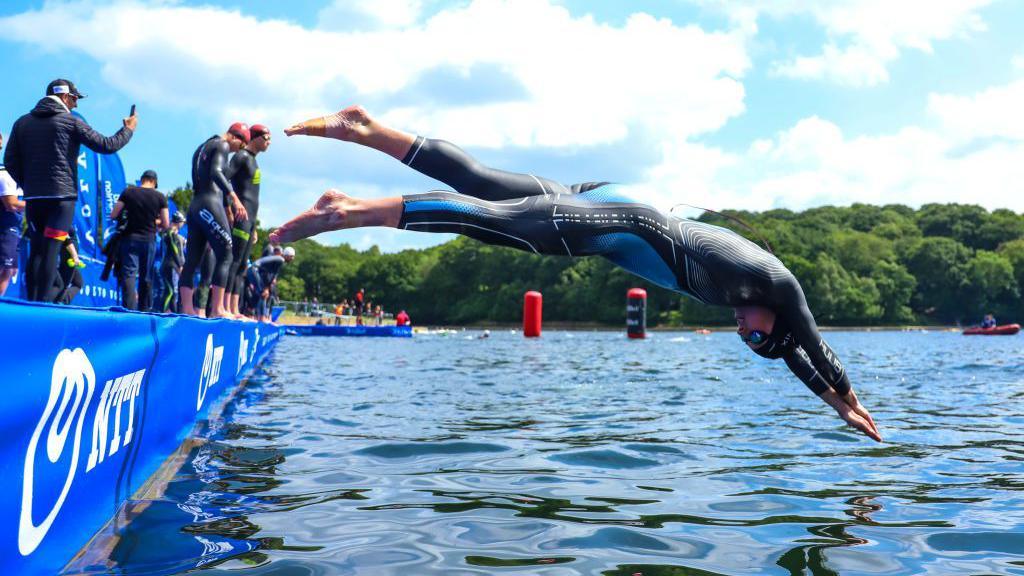 A man wearing a wetsuit dives off platform into a lake.