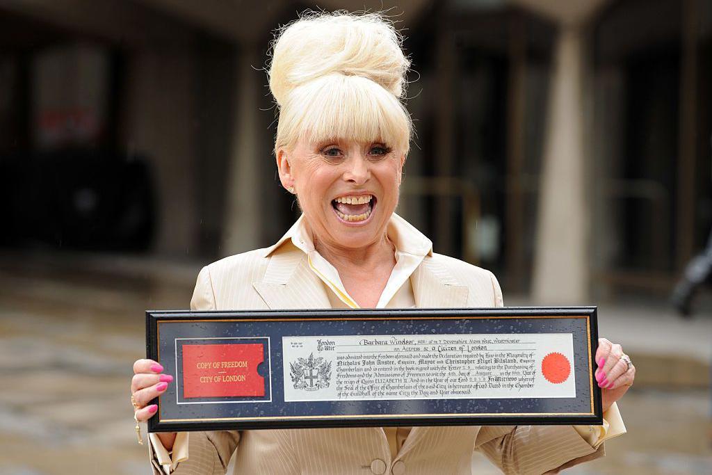 Barbara Windsor holds her Freedom of the City of London document. She is wearing a tan coloured jacket and has pink finger nails