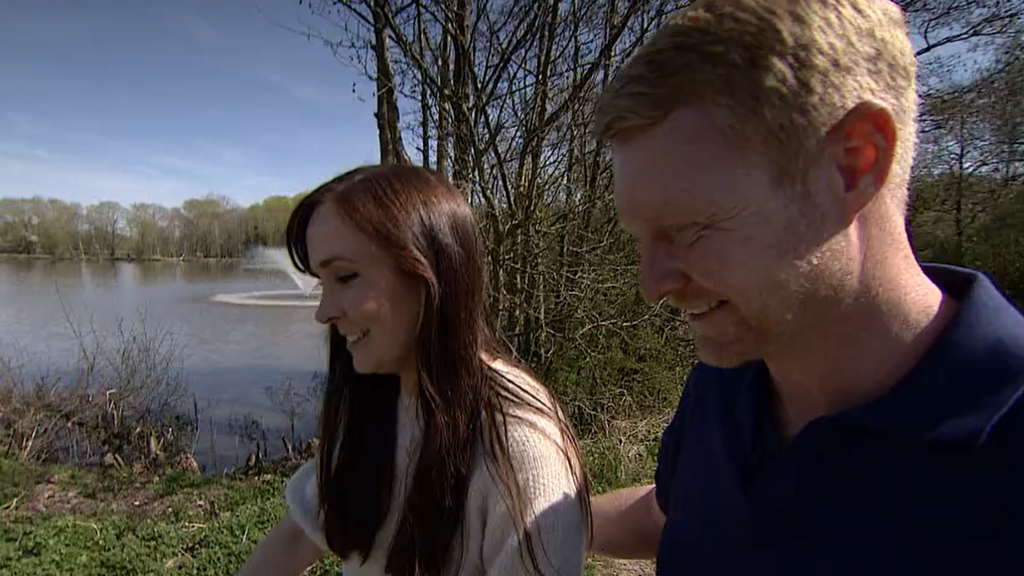 A smiling couple walk next to a lake with a water feature in the background. The woman has long dark hair and wears a light, cream top. The man to her left smiles and has short, red-blonde hair and stubble. He wears a blue top. Behind them is a grey-blue lake with trees around it.