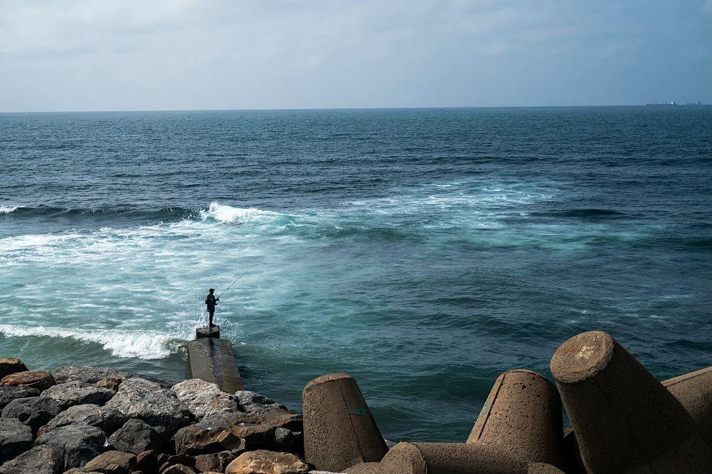 A man fishes from the shore. Behind him are large rocks and concrete wave breakers.