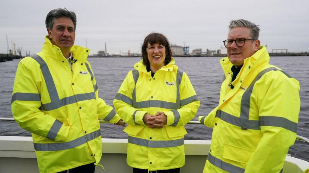 Ed Miliband, Rachel Reeves and Keir Starmer stand in high-vis yellow jackets with a backdrop of water behind them