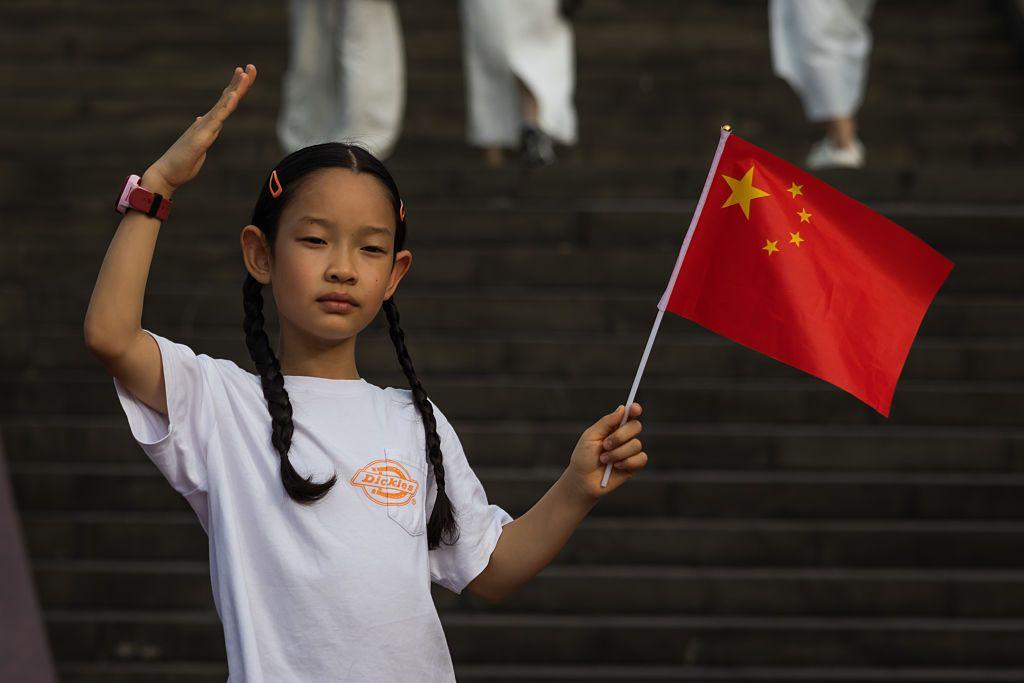 A girl waves a Chinese national flag and raises her hand in salute while standing on the steps near the Chongqing People's Auditorium during the National Day Golden Week holiday on October 7, 2025, in Chongqing, China