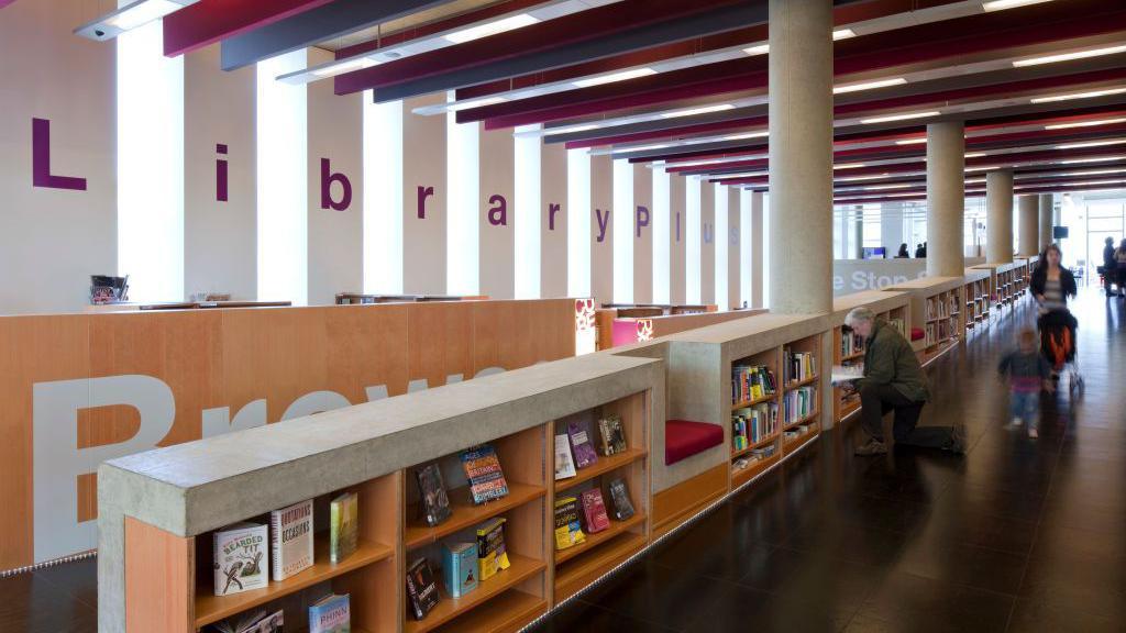 A library inside a modern building. The word library is written in purple on while pillars one letter at a time. There is a long row of short shelves with books. A man is on his knee reading a book while a woman with a pushchair and toddlers walk behind him.