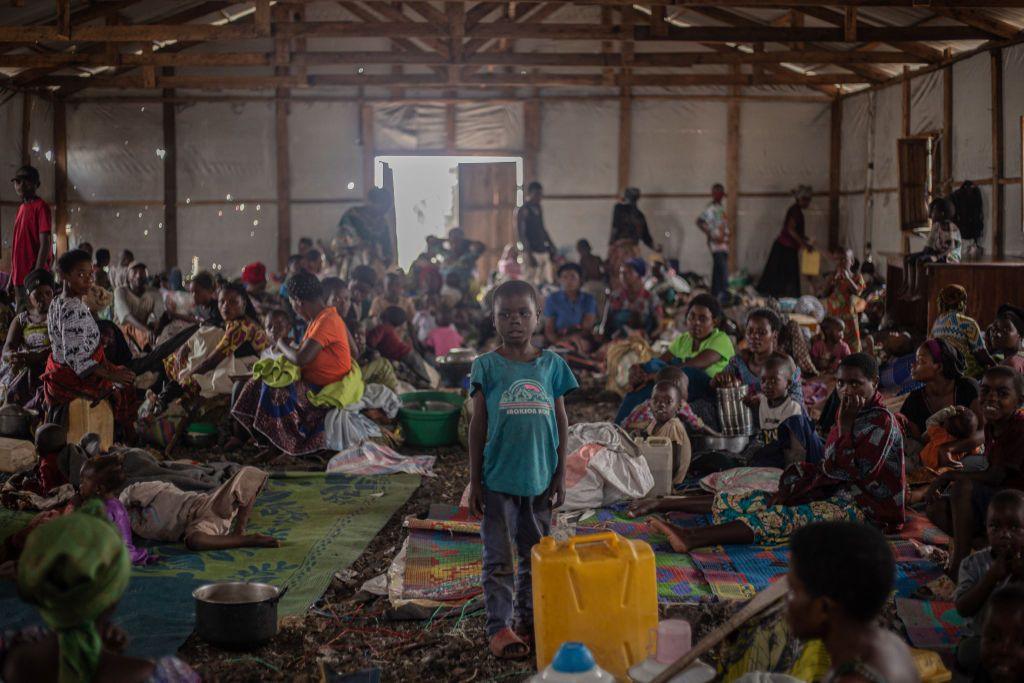 People stand crowded into a darkened makeshift home. They have very few belongings. Many are children.