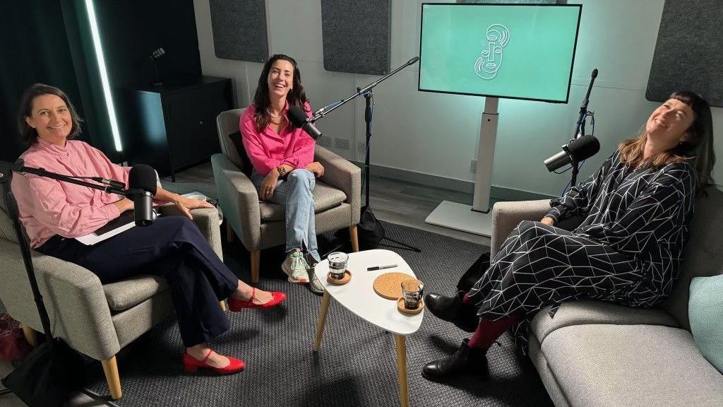 Three women sit on grey chairs with microphones on stands that are positioned next to their faces. They are smiling. There is a coffee table in the middle and a TV screen behind them