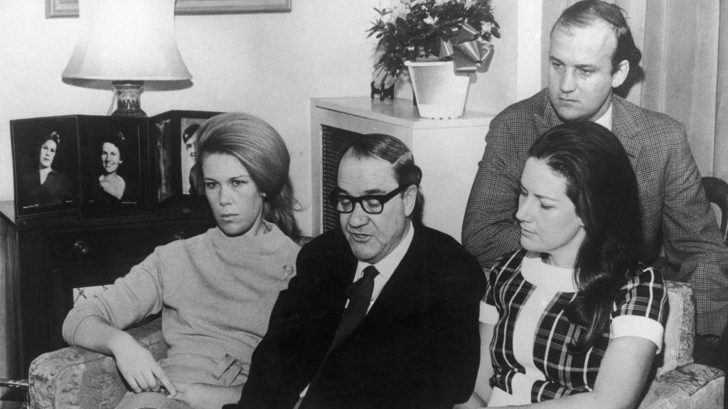 Alick McKay photographed with his children Dianne, Jennifer and Ian while reading out an appeal to her kidnappers for TV. They are sitting on a sofa in their living room and there is a photo of Muriel on the sideboard. Alick wears a dark suit and tie, while Jennifer wears a checked 1960s style dress, Ian wears a tweed jacket and Dianne wears a turtle necked sweater.