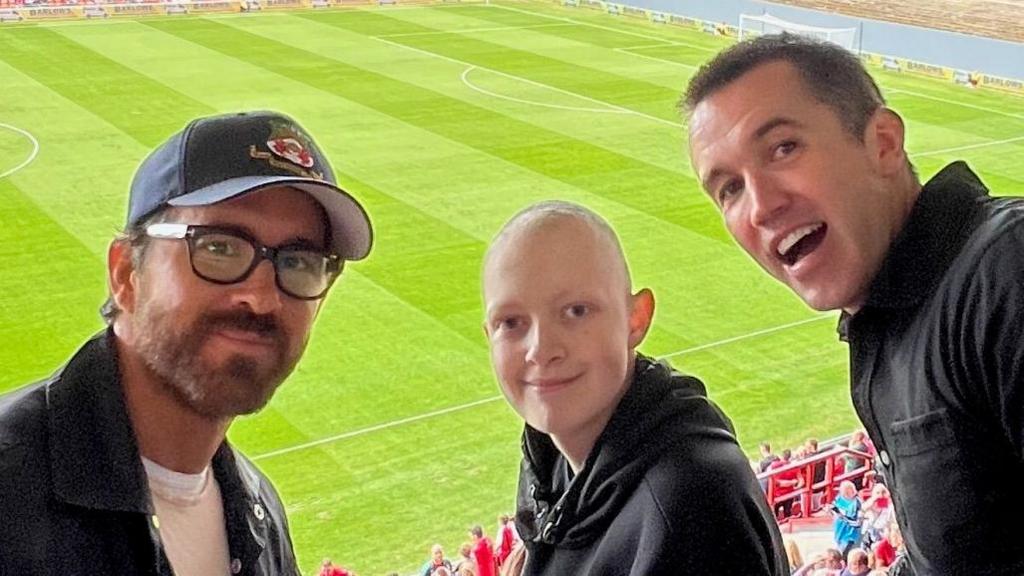 Reynolds, Aiden and McElhenney are pictured in the stands at Wrexham's stadium. All are smiling and the pitch is in the background.