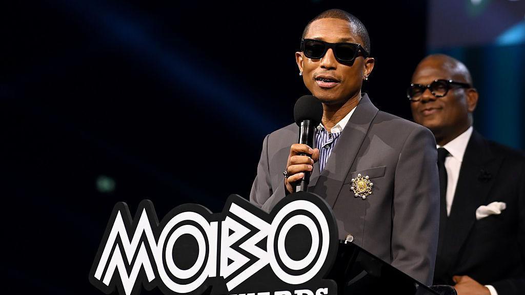 Harrell wearing a grey suit and black sunglasses talking into a microphone and standing in front of a podium that says Mobo 