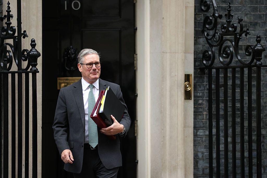 Sir Keir Starmer, a man with grey hair and glasses in a grey suit with white shirt and light green tie, is walking out of the front door of 10 Downing Street. The black door with gold numbering can be seen behind him. He is carrying a black and a maroon ring binder, both of which contain paper.