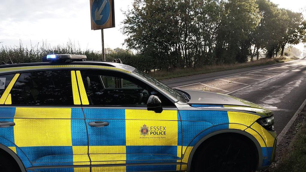 A close up shot of an Essex Police car which is parked diagonally across a road. The road has an area of white chevrons through the middle and is lined by trees. There is a keep left blue and white arrow sign behind the car, which is neon yellow and blue.