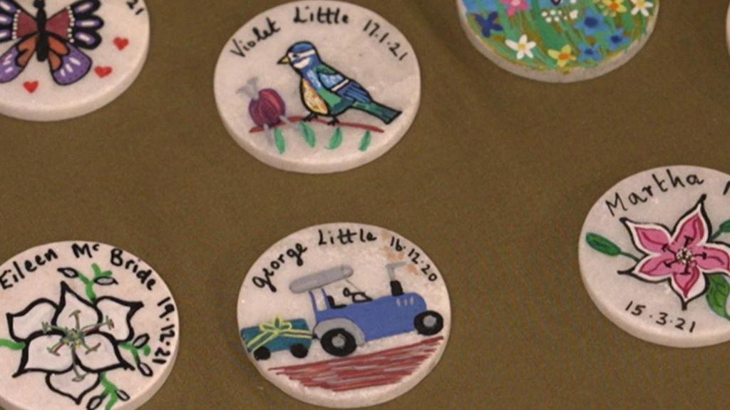 6 white badges with different designs sit on a brown table cloth. The badge on the bottom right is of a pink plant and the date 15/03/21. The badge in the middle has a blue tractor with the name and date, George Little, 16/03/20. Above that is one of a Blue tit and a pink ball of wool with knitting needles, it says Violet Little, 17/01/21. Bottom left is of white flowers and the name Eileen McBride, 19/12/21. Top left has a drawing of a purple and orange butterfly. Top right is of a garden filled with a variety of flowers. 