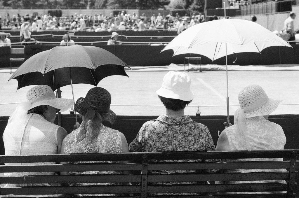 Black and white image showing four ladies on a courtside bench shading with umbrellas as they watch the tennis at Wimbledon.