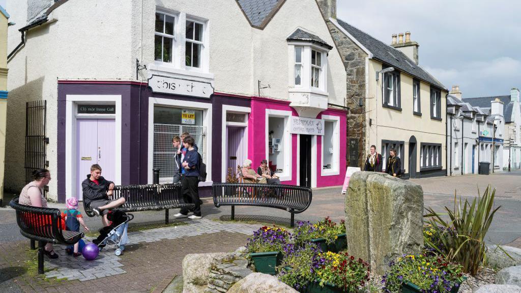 A generic photo of Stornoway on the Isle of Lewis, you can see some people sitting on a black bench and some brightly coloured shop fronts in the foreground