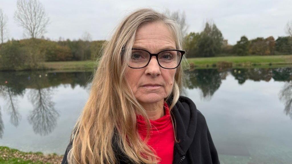 A woman with long blond hair and black-rimmed glasses looks towards the camera. In the background is a lake with some trees dotted around.