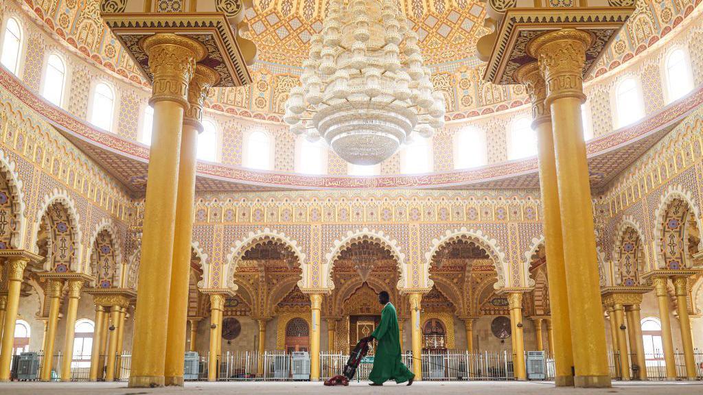 A man in a green tunic runs a vacuum cleaner over the mosque's carpet. The mosque's opulent-looking pillars, walls and ceiling are in shot - in Dakar, Senegal, Thursday 27 February 2025.