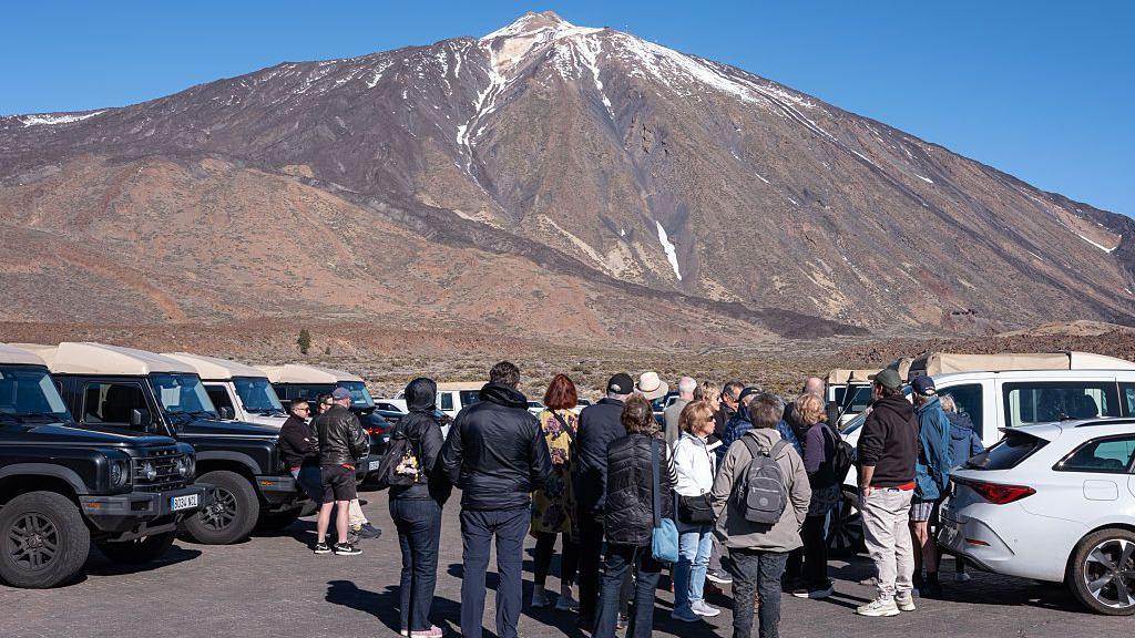 The peak of Mount Teide, with a little snow on its sides, bathed in sunshine with a blue sky overhead, with a car park full of cars and people in the foreground.