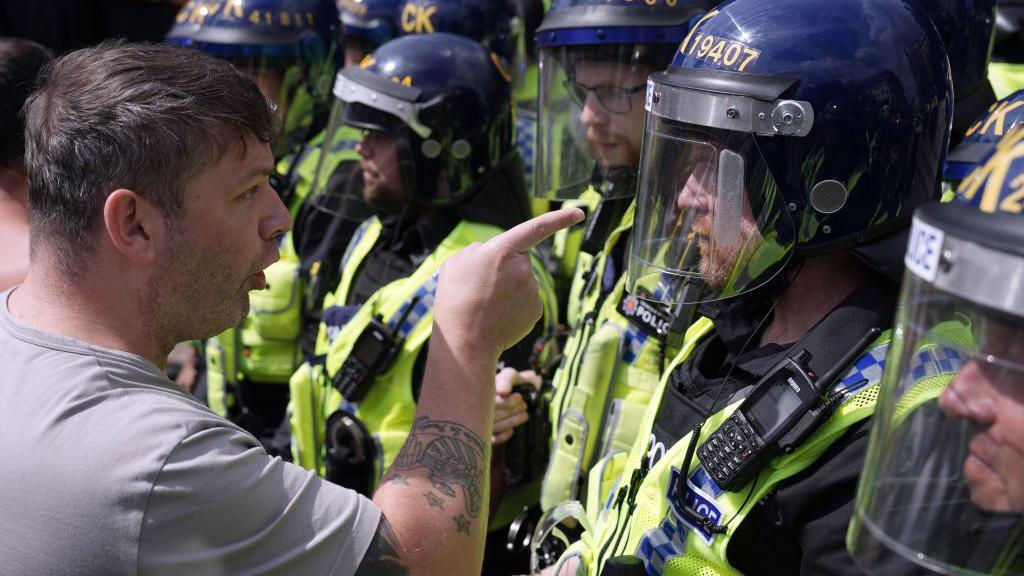 A man with short head and tattoos points his finger in the faced of a police officer. The man appears to be saying things to the officer, who appears to be looking away from him.