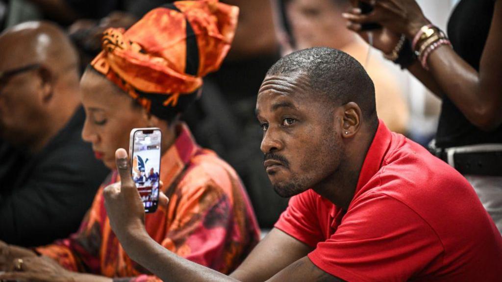 The leader of the Rally for the Protection of Afro-Caribbean Peoples and Resources (RPPRAC) Rodrigue Petito, wearing a red T-shirt, looks at his phone, during what appears to be a meeting. 