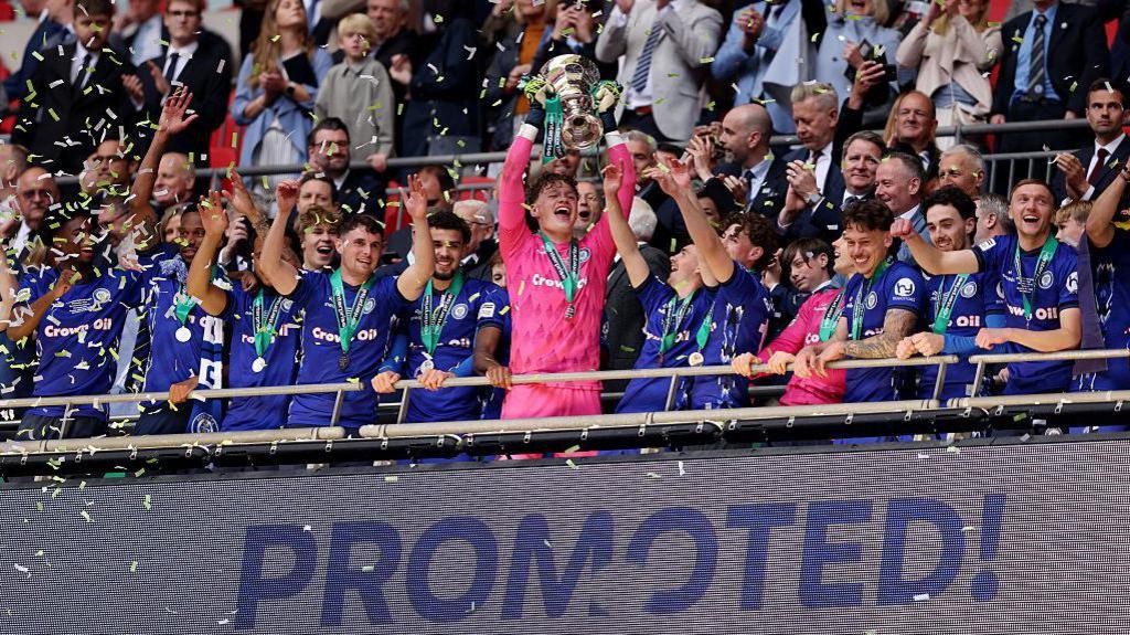 Oli Whatmuff lifts the National League promotion final trophy above a sign saying 'promoted'