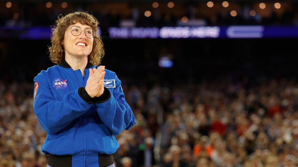 Christina Koch wearing a blue nasa jacket with her hands clasped together in front of a big crowd that's out of focus