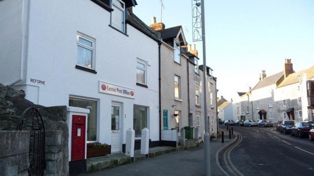 An old photo of the post office in Reforne, Easton. There is a red wall post box outside the whitewashed terraced building. The rest of the houses in the street are grey or bare stone.