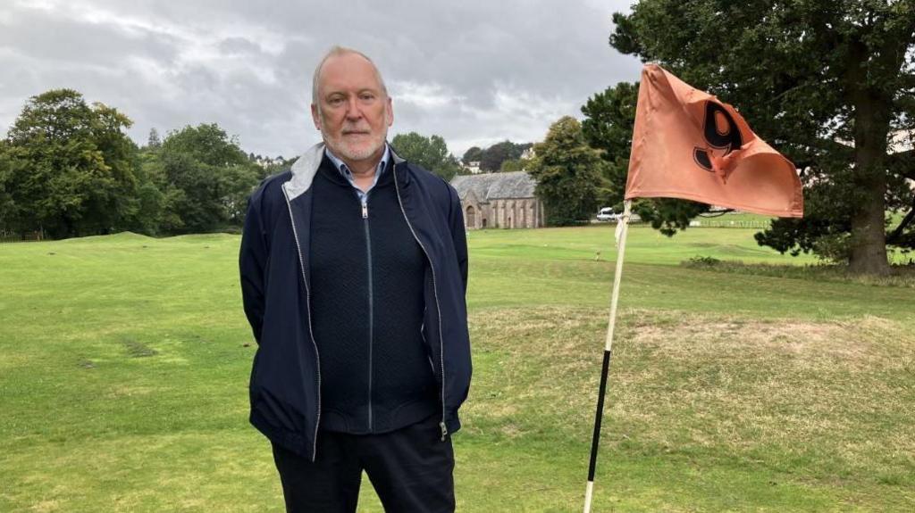 Anthony Mills wearing a blue coat over a navy blue fleece stood on a golf course next to a orange flag with a number nine on it. He is stood about 100 metres away from the stone-built Spanish Barn.