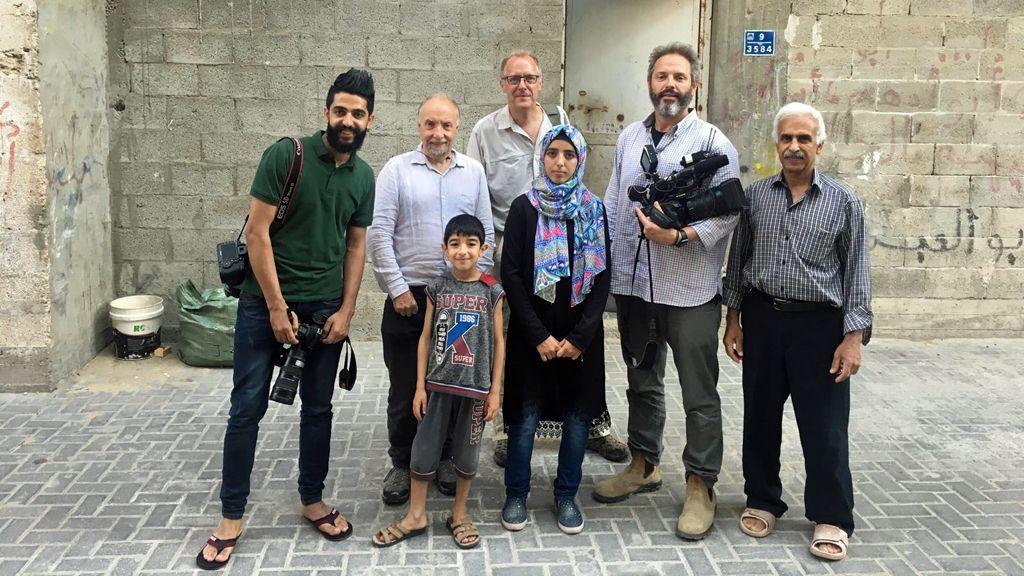 Asmaa, a petite woman in a colourful headscarf, pictured in the middle of a group of five men and a small boy. One of the men is holding a video camera. Paul Adams, a tall man with glasses, is standing behind her. They are pictured in a concrete courtyard and there is some grafitti on the walls. The door to her home is behind them.