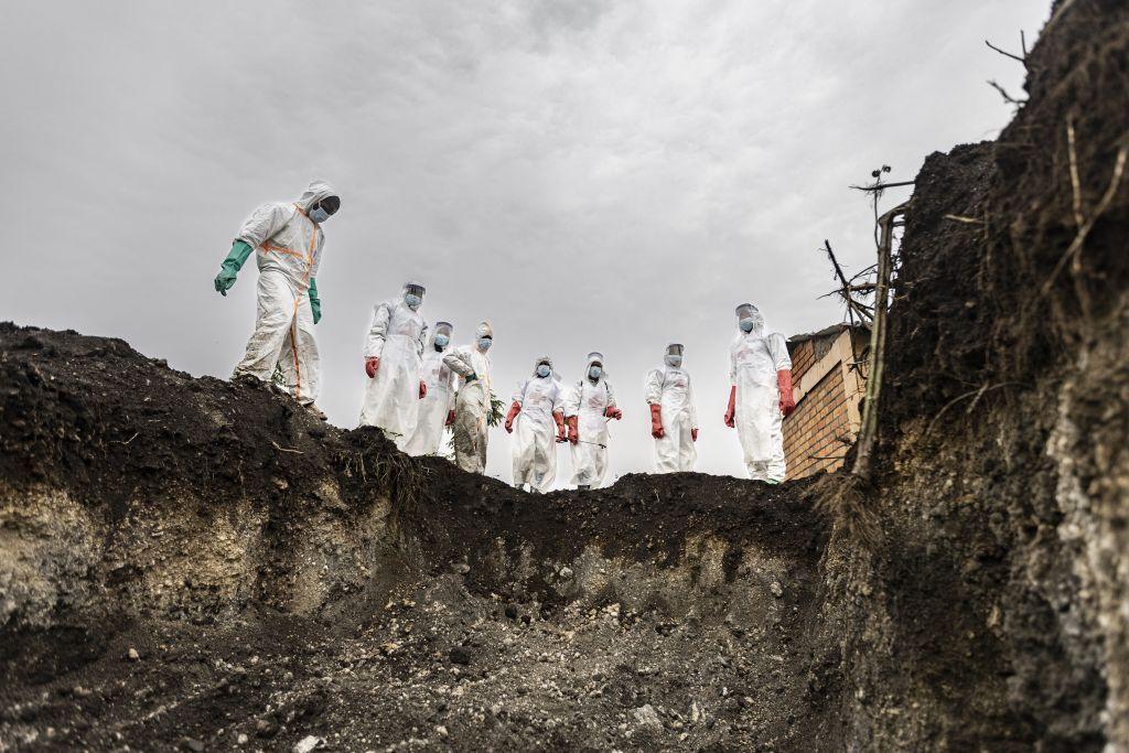 Undertakers line up at the edge of a dug mass grave. It is empty, ready for bodies to be laid to rest in it.