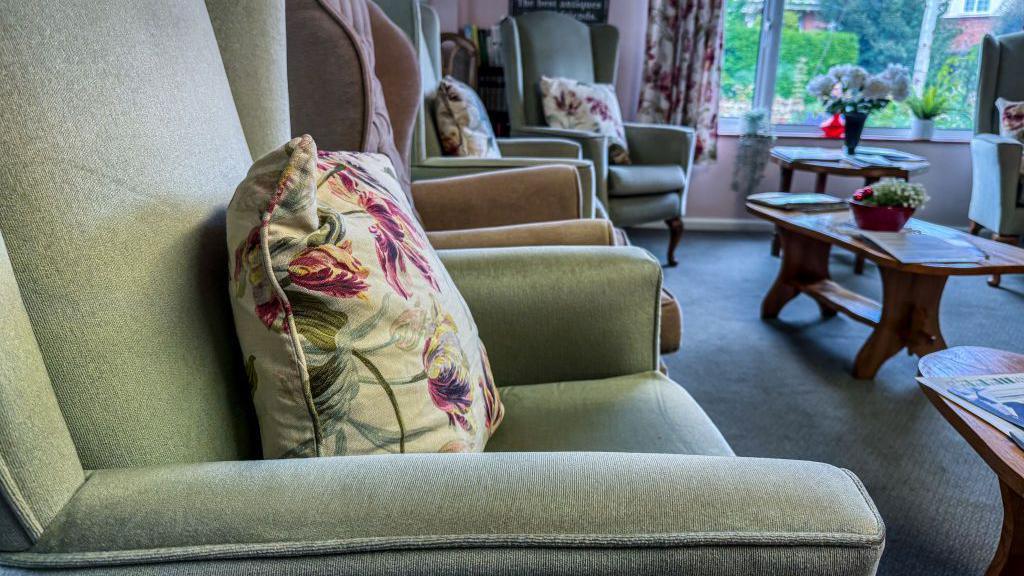 The interior of a empty care home living room with four upright chairs with cushions on and three wooden tables.