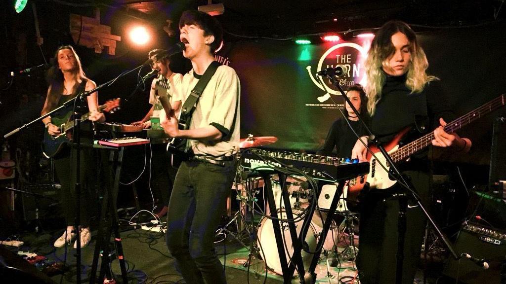 Declan Mckenna is on stage with his band singing into a microphone and playing guitar