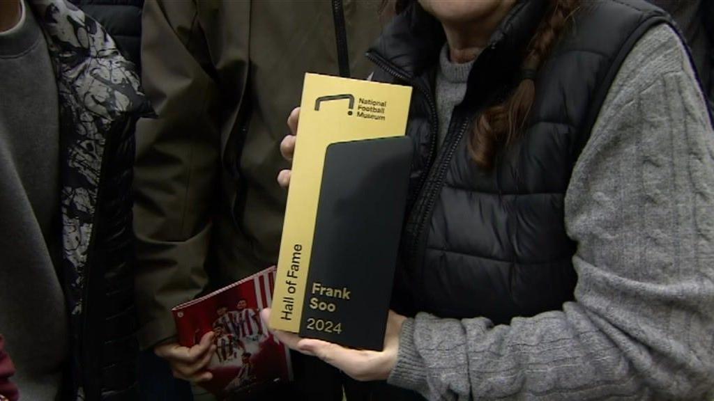 A close-up shot of a woman stood among a group of people, as she holds a black and gold plaque which has the name of Frank Soo engraved on it as well as the National Football Museum logo.