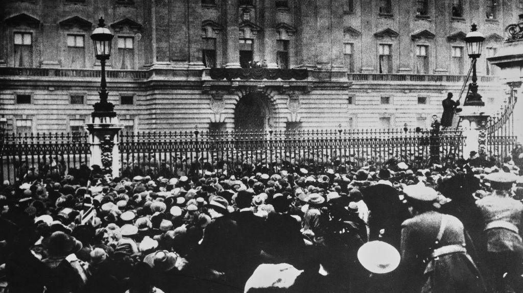 Black and white photo showing a crowd outside the gate of Buckingham Palace on 11th November 1918. In the background George V and members of the Royal family can be seen on the balcony of the palace.