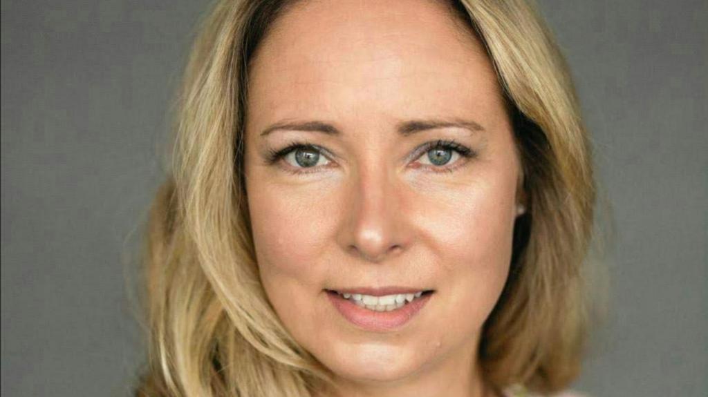 Catherine McKeown pictured with long blonde hair. She is smiling and standing in front of a grey screen (for a company photo)