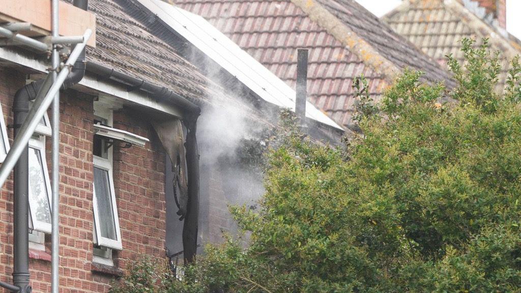 A charred building with smoke coming out of it. A green bush is in the foreground, shielding the house from view.