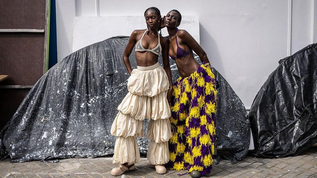 Two models stand in front on a street posing. They're wearing frilly trouser bottoms.