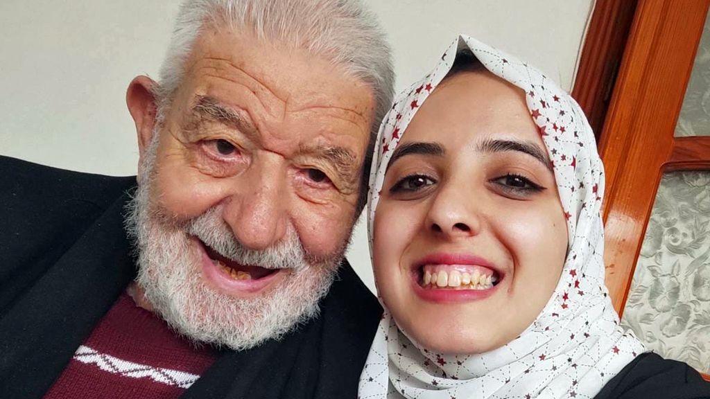 A selfie of Asmaa, wearing a white headscarf patterned with stars, smiling, wearing winged eyeliner, with her grandad, who has a white beard. He is leaning towards her and his head is touching hers as they look to the camera.