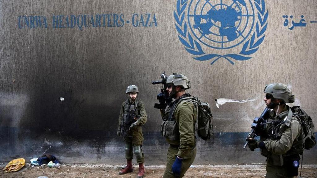 Israeli troops in front of a wall bearing the Unrwa logo