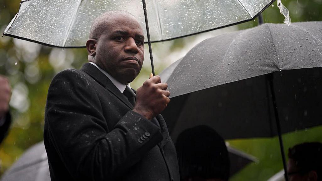 David Lammy stands under a clear plastic umbrella and is looking to his right into camera. He is wearing a black raincoat, white shirt and black tie and looks very serious. There are many raindrops on the umbrella. 