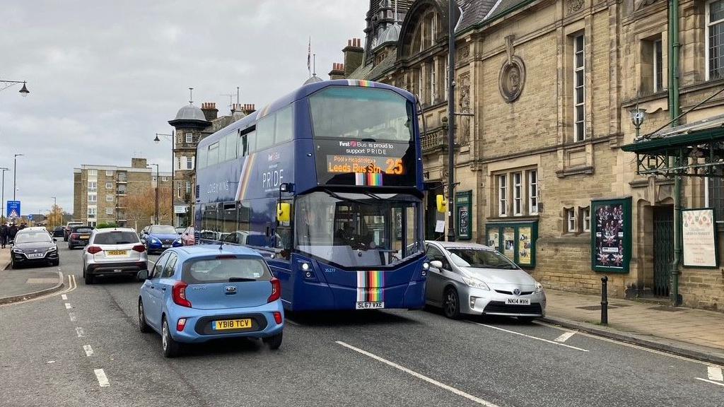 A double decker bus in blue livery driving in between two cars going in the opposite direction on a road in Ilkley. 