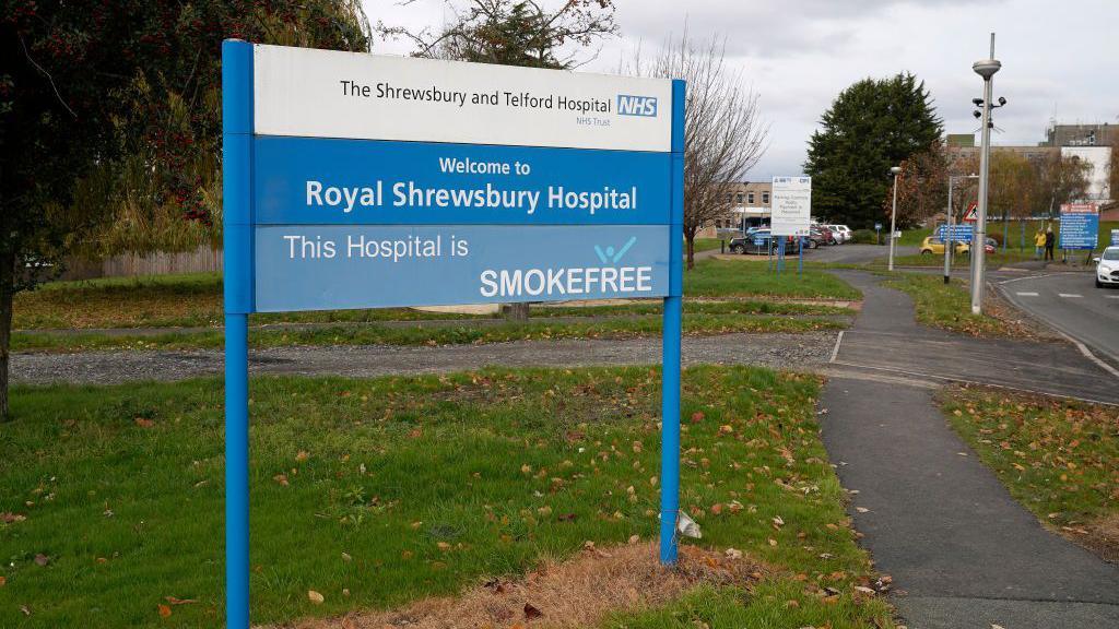 A blue and white NHS sign at the entrance to the Royal Shrewsbury Hospital. There is a car park in the background. 