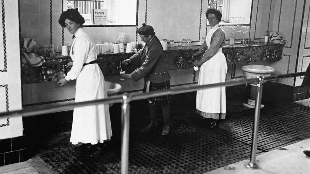 Two women dressed in Victorian clothing and a boy wearing a kilt fill cups with water from taps.