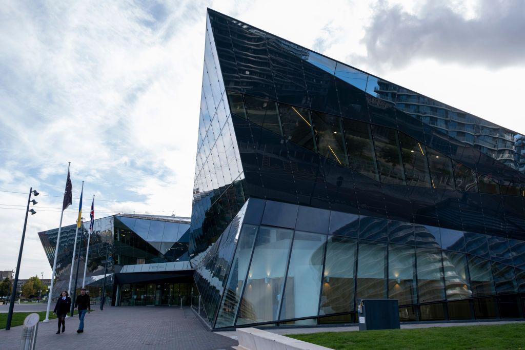 The exterior of the London City Hall building in Newham. A large, dark glass building is shown, with a walkway leading to the entrance.