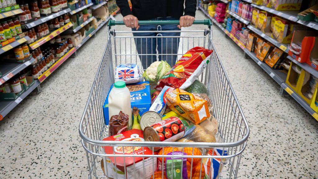 a person pushing a trolly with various food items in it like milk, crackers, tinned tomatoes and cereal