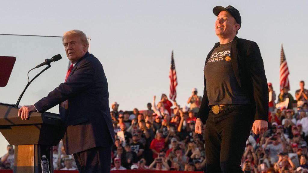 Donald Trump, in a navy suit and red tie, stands at a podium, looking sideways at the camera while Elon Musk, dressed in black, beams before a crowd a with American flags in the background