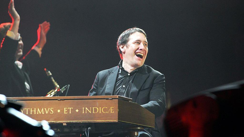 Jools Holland wearing a black suit laughing on stage sat at his piano