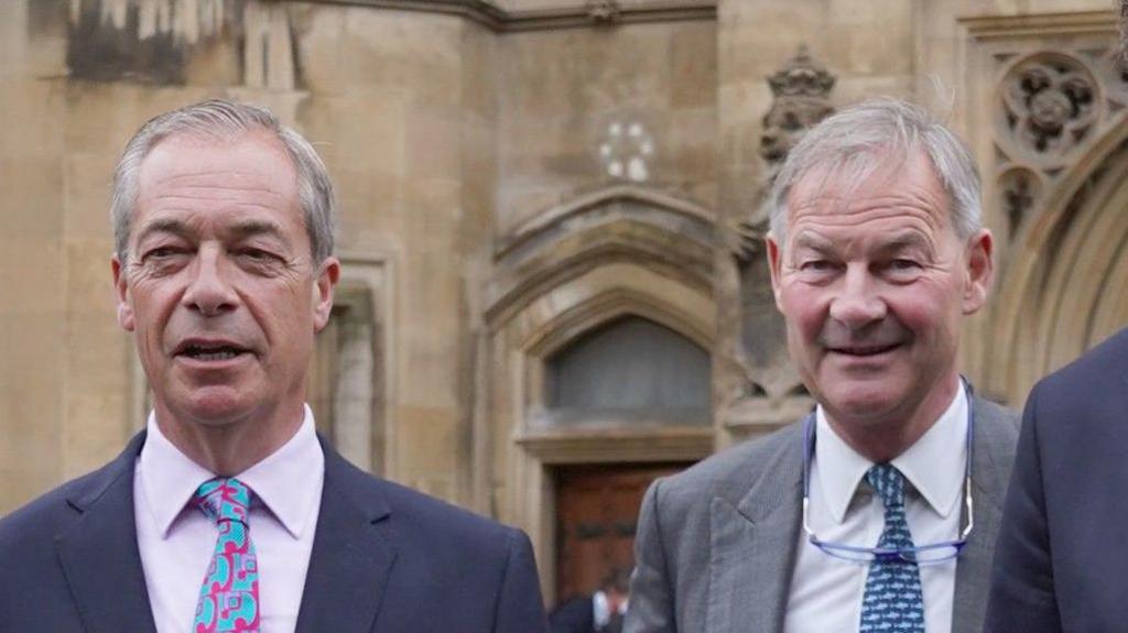 Nigel Farage and Rupert Lowe standing next to each other. They are both wearing suits with shirts and ties. 