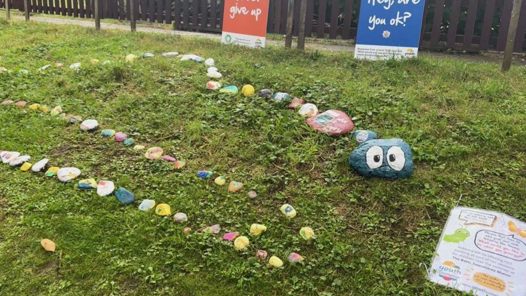 Colourful painted stones forming the snaking body of a caterpillar, with a large blue stone acting as its head, all on grass