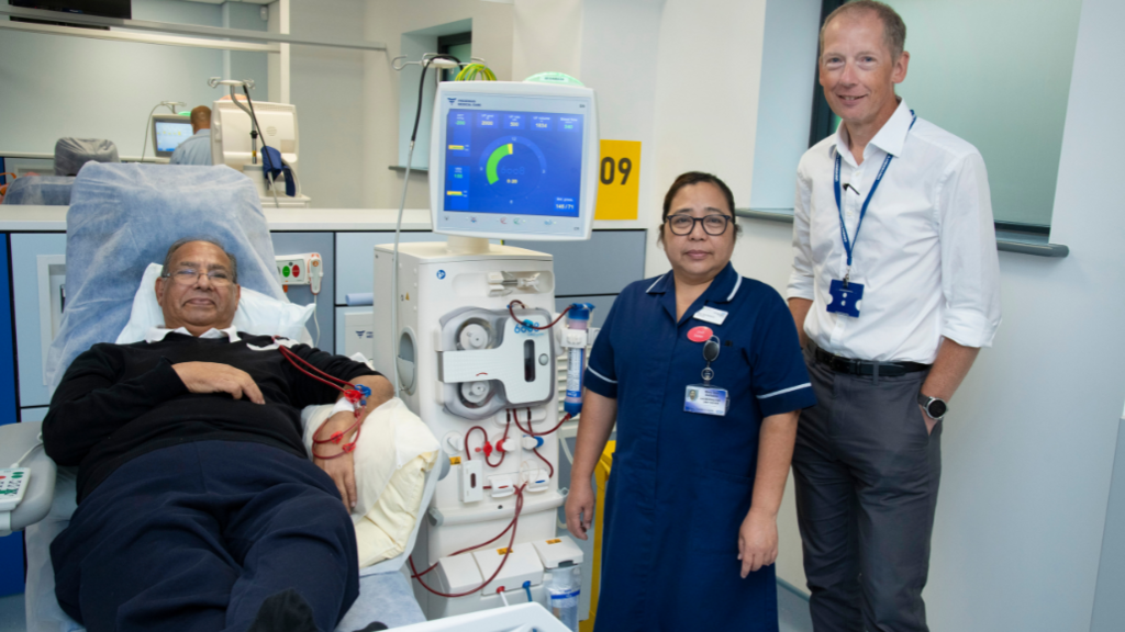 On the left, a man is lying on a hospital bed with wires in his arms that are attached to a dialysis machine. On the right, is a female member of staff wearing a navy blue nurse's uniform, stood next to a male staff member in a white shirt and grey trousers 