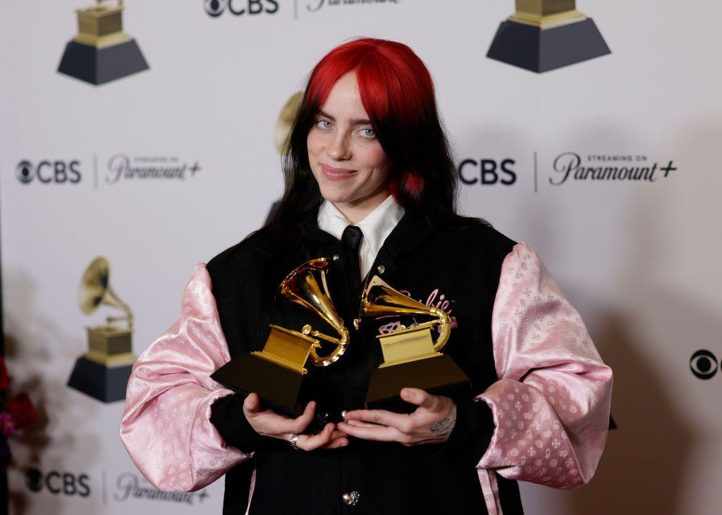 Billie Eilish holds up two Grammy Award trophies, she is smiling and is wearing a pink and black jacket