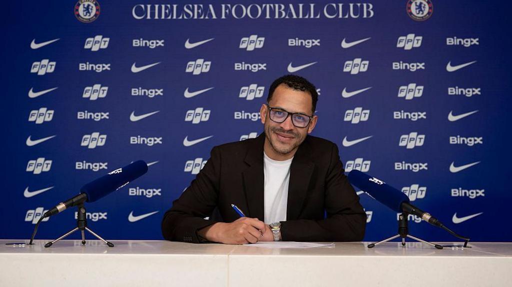 Photo showing a smiling Liam Rosenior signing his Chelsea contract, with advertising logos behind him on a wall also bearing the words Chelsea Football Club and two club badges
